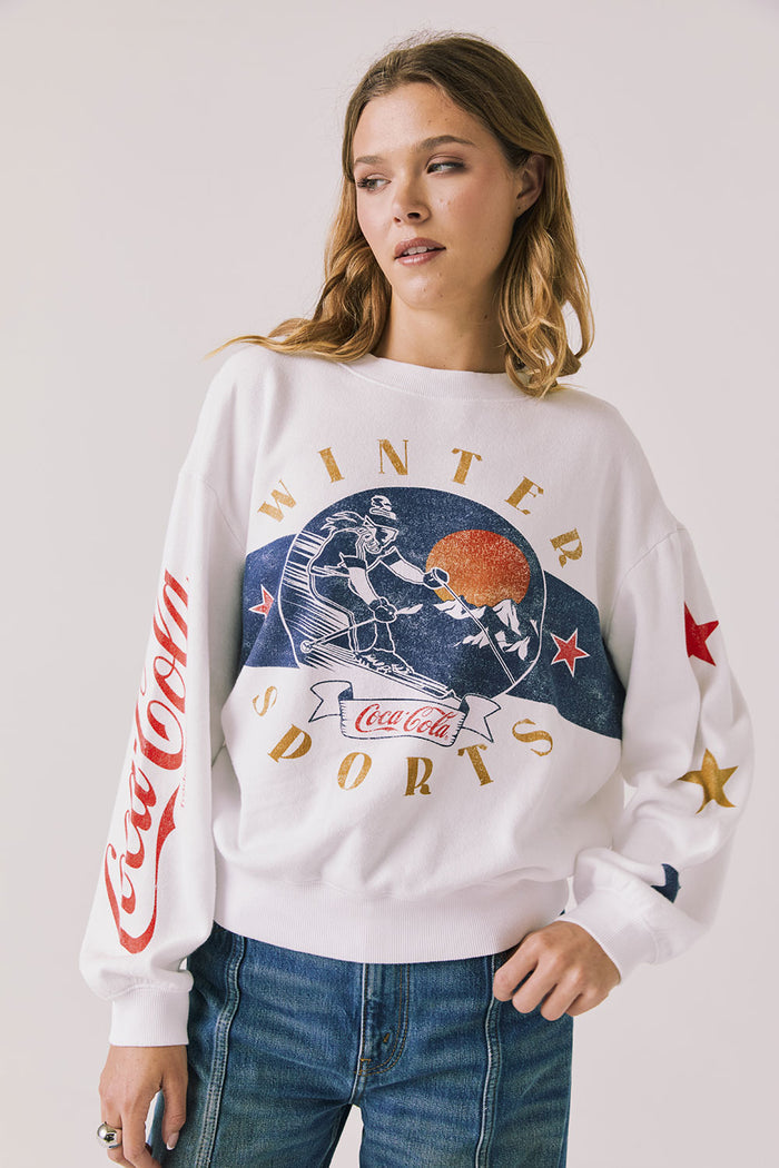 Front view of woman wearing Chaser’s white Coca-Cola Winter Sports Long Sleeve with vintage-inspired graphic design and blue jeans on a plain background