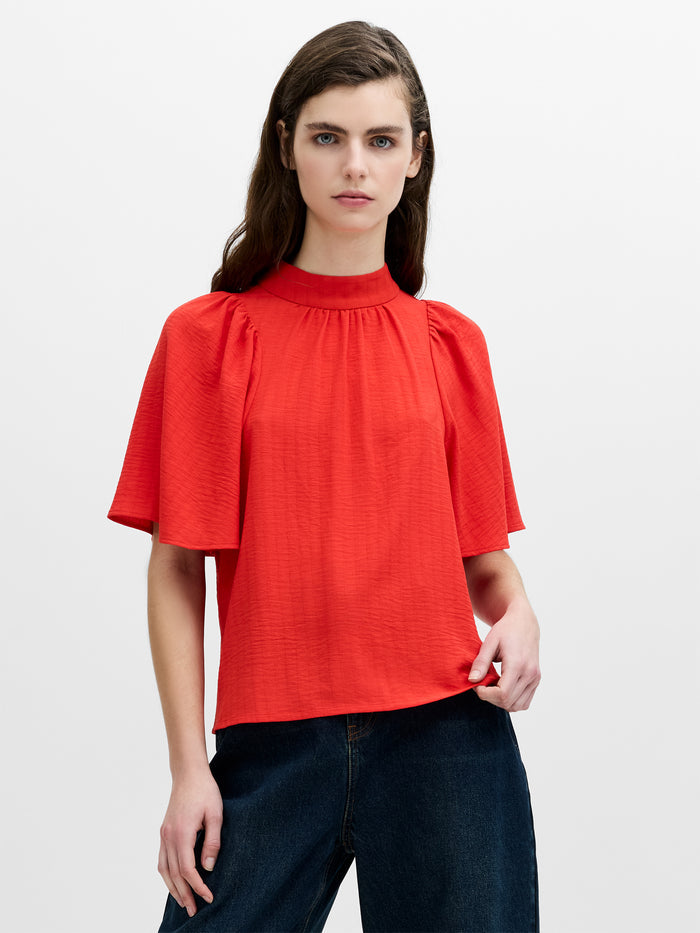 A woman wearing a red blouse with short flowy sleeves and a high neckline.