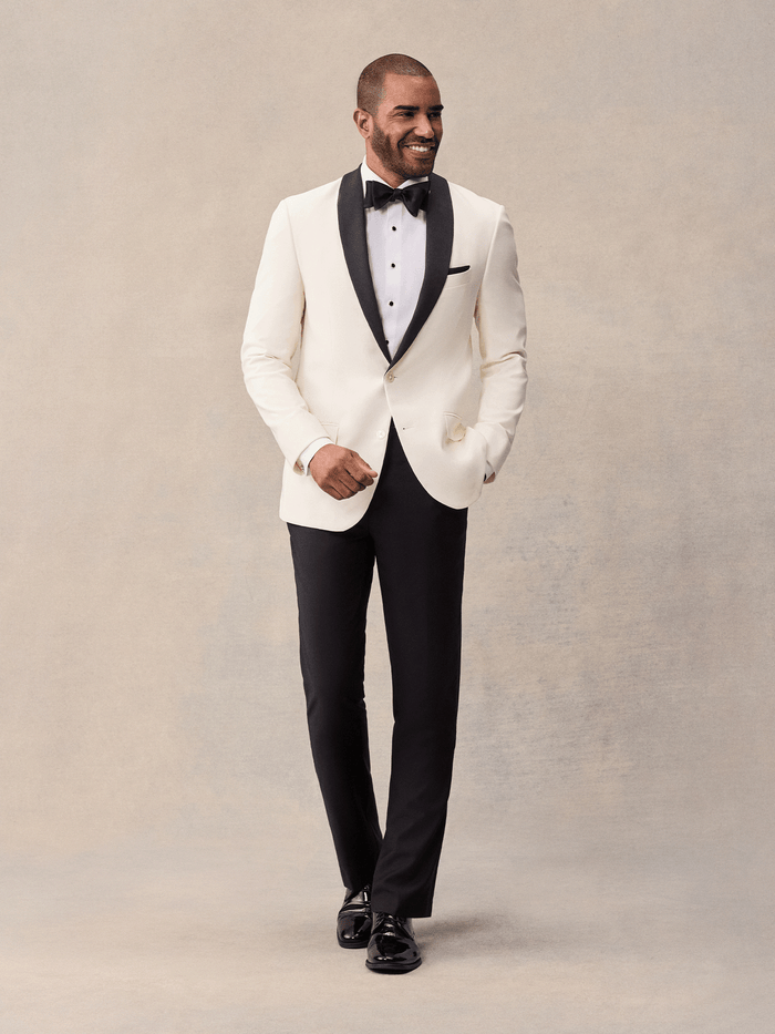Man walking in an ivory shawl-lapel tuxedo jacket with black lapels and black trousers, full length.