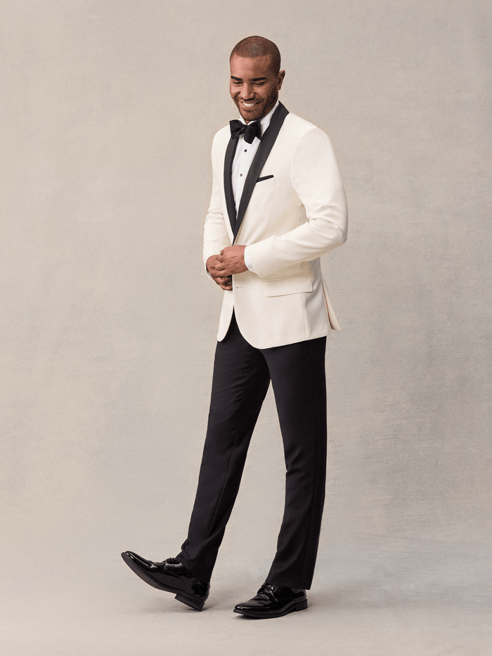 Man in an ivory shawl-lapel tuxedo jacket shown at a three-quarter angle, hands clasped.