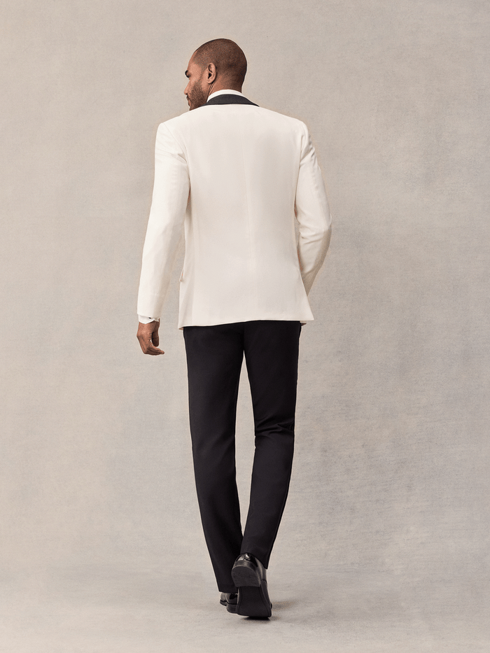 Back view of a man walking in an ivory shawl-lapel tuxedo jacket and black trousers.