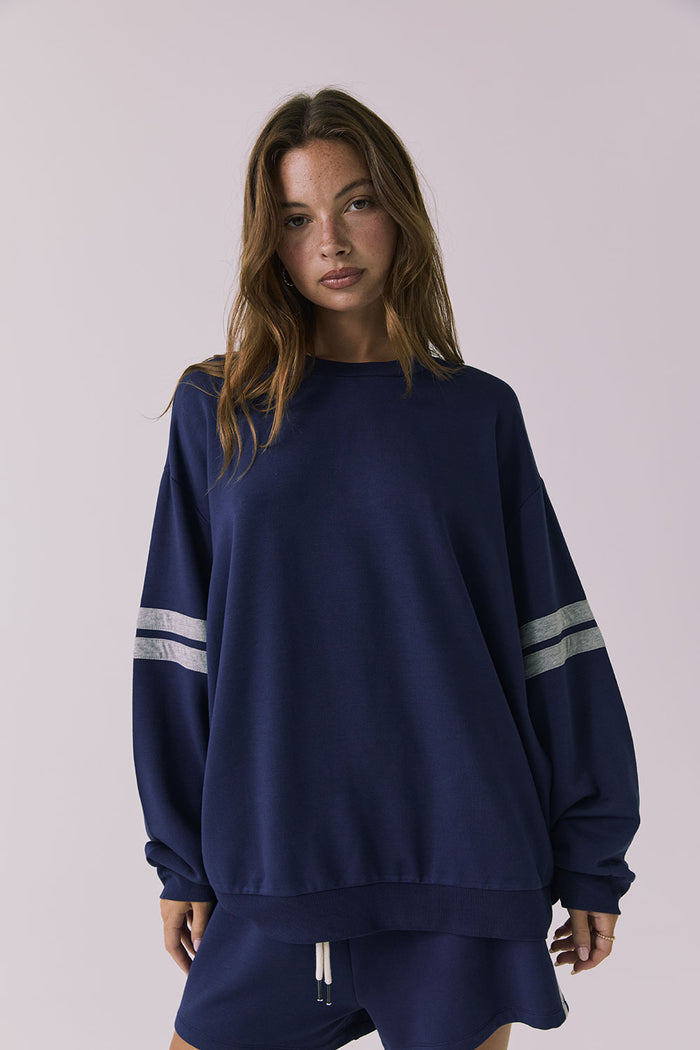 Front view of a woman wearing Chaser's navy blue Cornell Sapphire & Heather Grey Pullover with grey stripes on sleeves against a light gray background