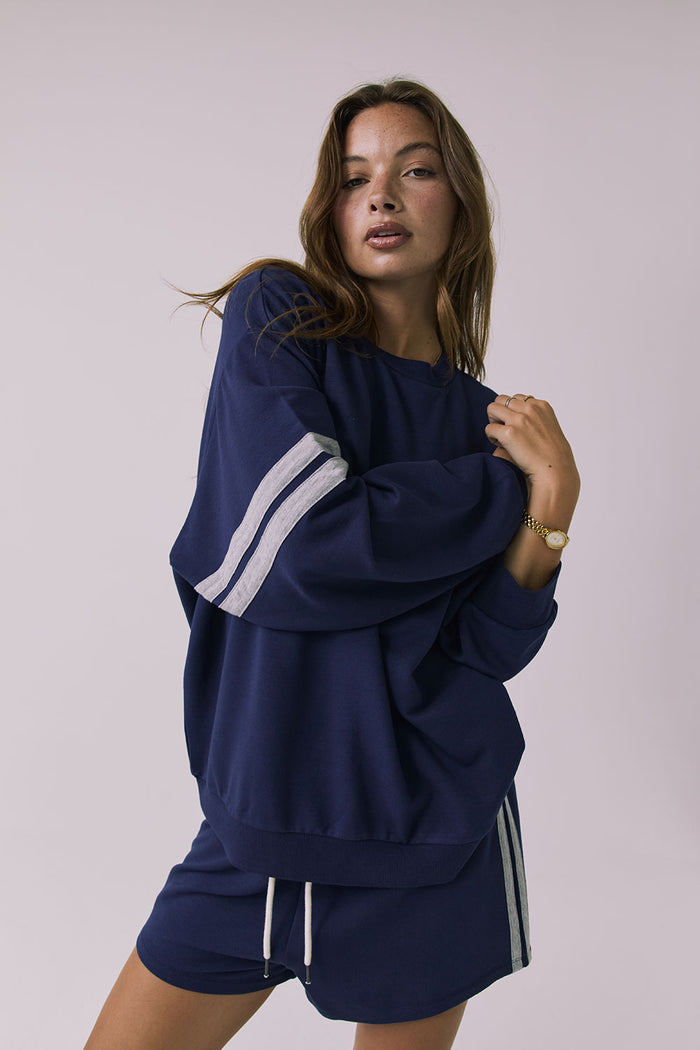 3/4 view of a woman wearing Chaser's navy blue Cornell Sapphire & Heather Grey Pullover with grey stripes on sleeves against a light gray background