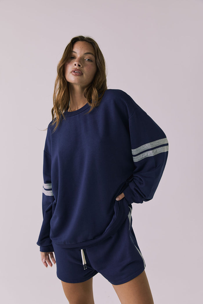 Front view of a woman wearing Chaser's navy blue Cornell Sapphire & Heather Grey Pullover with grey stripes on sleeves and matching Hanoverr shorts on a plain background