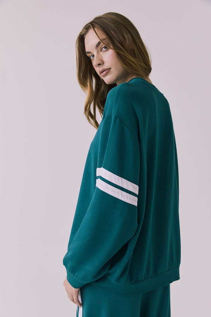 Back view of a woman wearing Chaser's Cornell Pullover in Teal and White with striped sleeves on a plain studio background