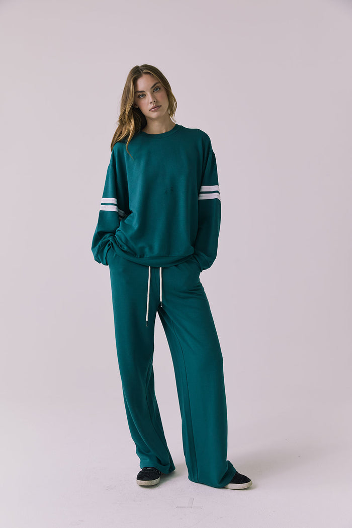Full body front view of a woman wearing Chaser's Cornell Pullover in Teal and White with striped sleeves and the matching Sandi Joggers on a plain studio background