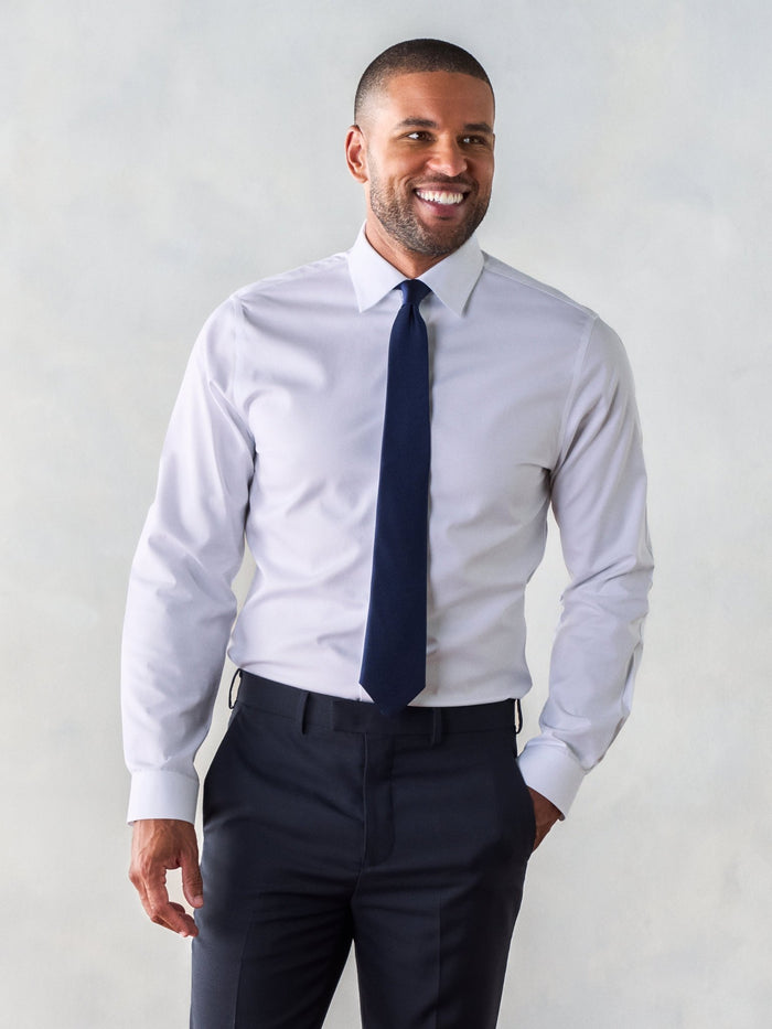 Man in a white dress shirt with a navy tie and dark trousers, hand in pocket.