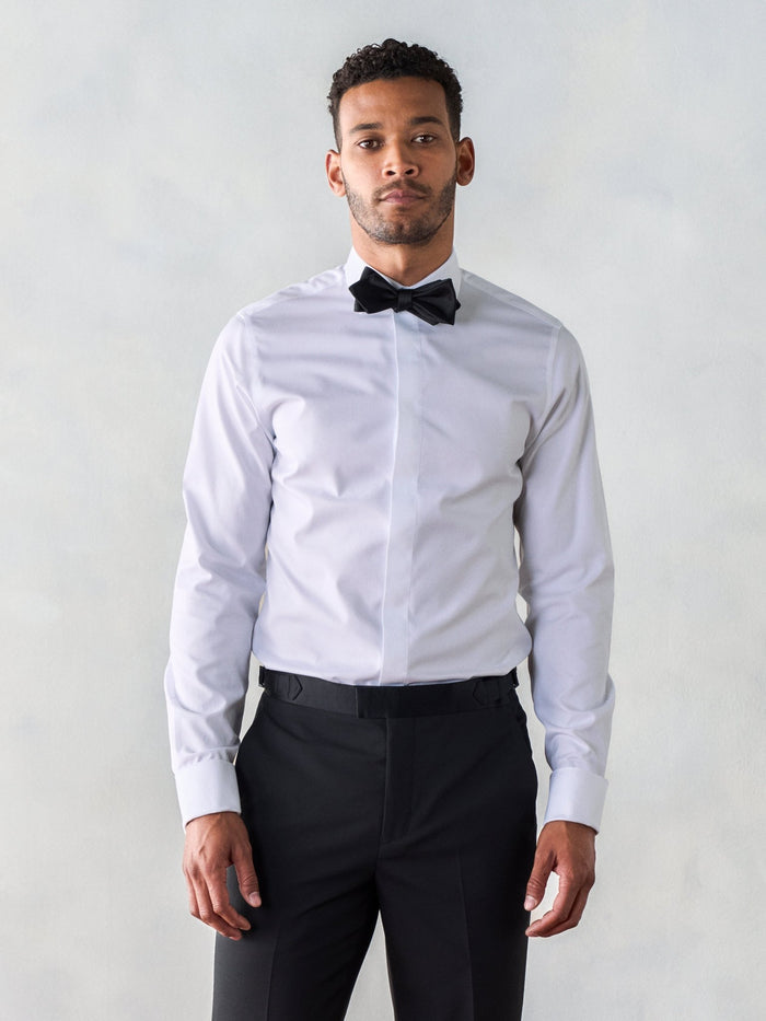 Man in a white fly-front tuxedo shirt with a black bow tie and black trousers.