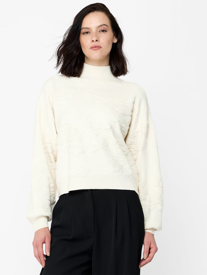 A woman wearing a cream knit Jumper with a floral pattern, long balloon sleeves and a ribbed mock neckline.