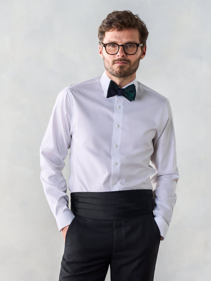 Man in a white tuxedo shirt with a black pleated cummerbund and dark green plaid bow tie.