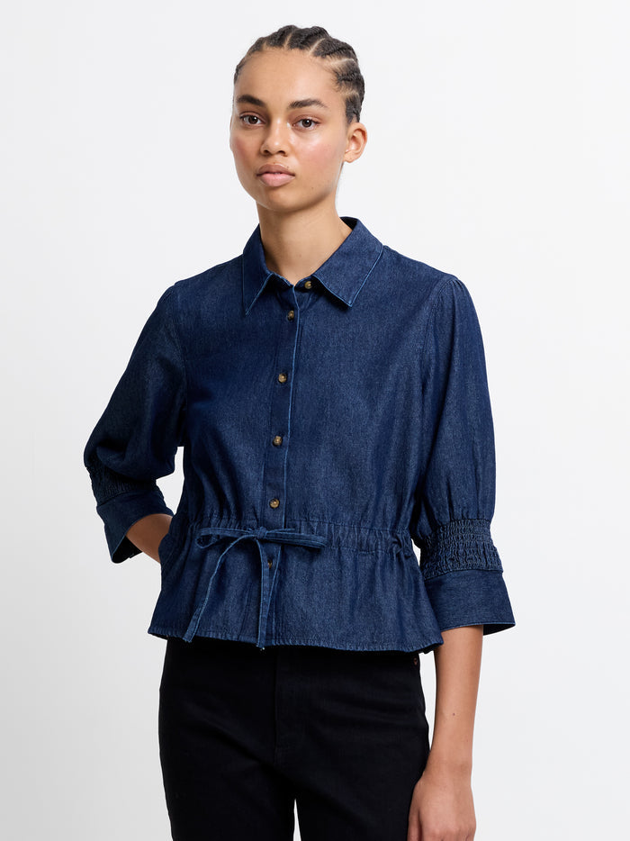 A woman wearing a dark blue smocked sleeve self-tie drawstring top with mid length sleeves, peplum hem and a button down fastening.