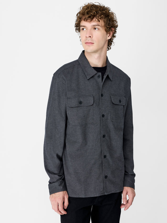 A man wearing a grey shirt with long sleeves, 2 front pockets, a pointed collar and button down fastening.