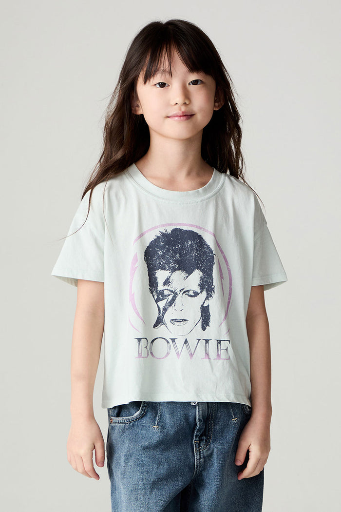 Side view of a girl wearing the Chaser David Bowie Stars Bowie Girls Tee on a neutral background with bluejeans and arms at her sides
