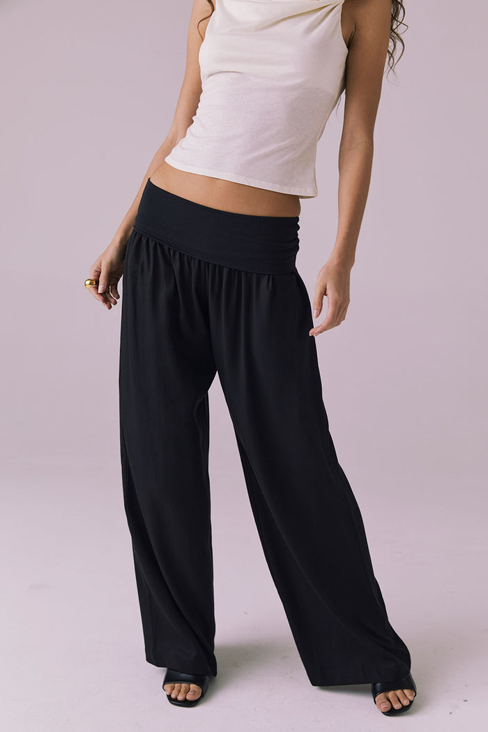 Model wearing the Chaser Didion Licorice Black Trousers leaning slightly forward to show the soft drape and foldover waistband