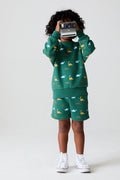 Full body view of a boy wearing Chaser Kids Dino Party Boys Shorts in green with matching Dino Party Pullover, holding a camera, with white shoes against a plain background