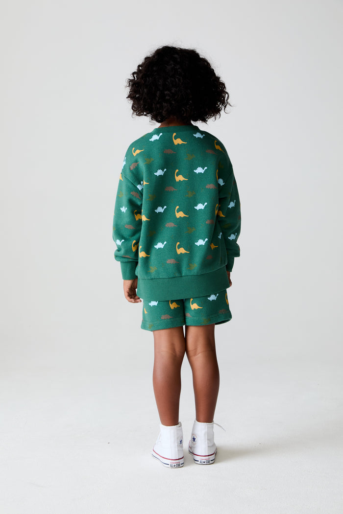 Back body view of a boy wearing Chaser Kids Dino Party Boys Shorts in green with matching Dino Party Pullover with white shoes against a plain background