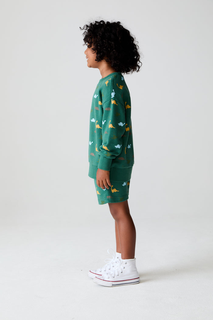 Side body view of a boy wearing Chaser Kids Dino Party Boys Shorts in green with matching Dino Party Pullover with white shoes against a plain background