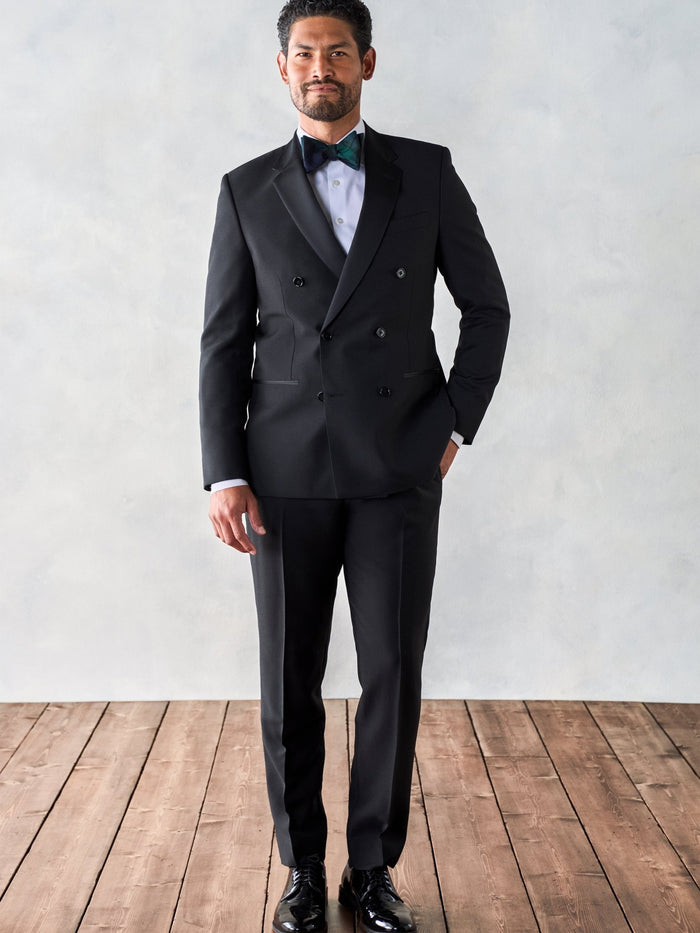 Man in a black double-breasted tuxedo, full length standing on a wood floor.