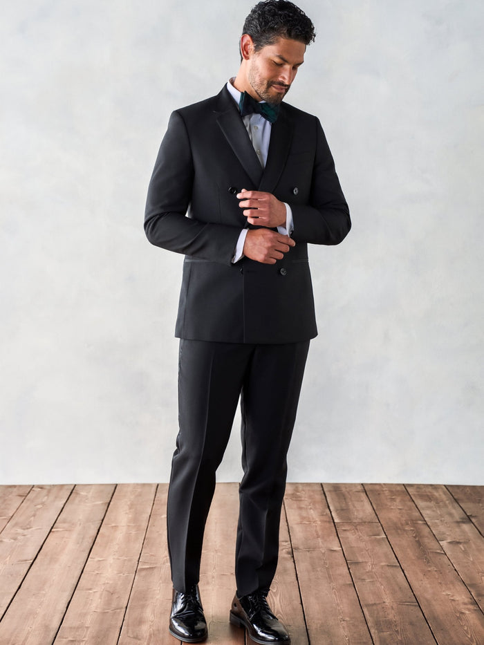 Man buttoning a black double-breasted tuxedo jacket, full length, on a wood floor.