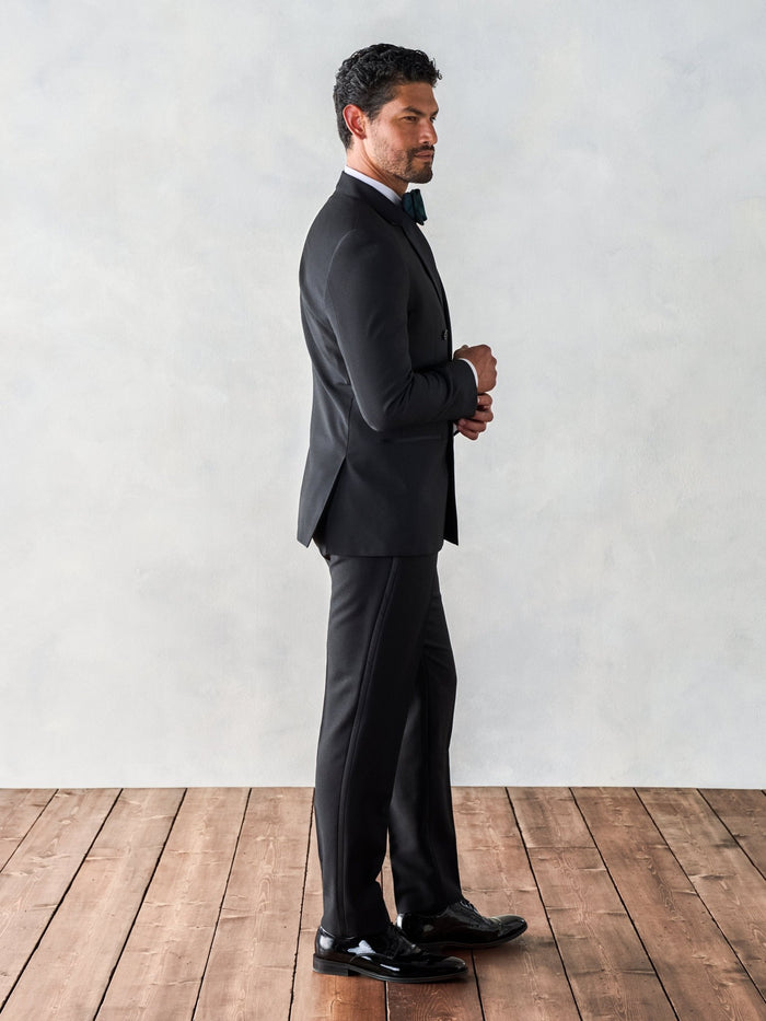 Side view of a man in a black double-breasted tuxedo, full length on a wood floor.