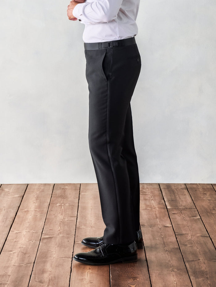 Side view of a man from waist down in black tuxedo trousers, standing on a wood floor.