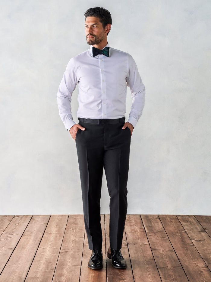 Man in a white dress shirt, patterned bow tie, and black trousers, standing on a wood floor.
