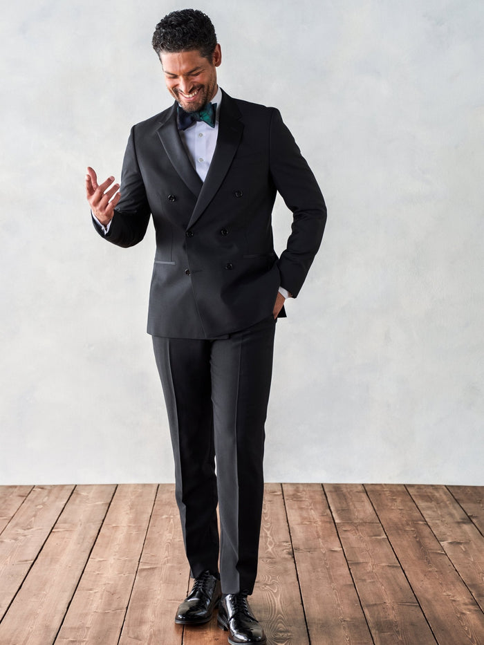 Man in a black double-breasted tuxedo smiling and looking at his hand on a wood floor.