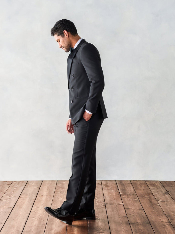 Side view of a man in a black double-breasted tuxedo looking down at a wood floor.