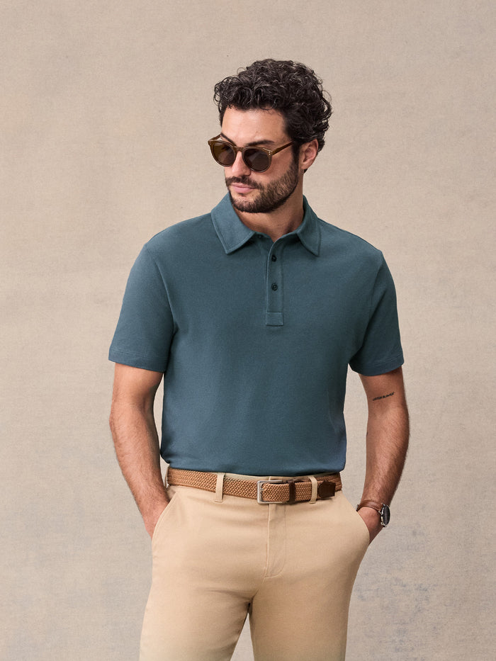 Man in slate blue pique polo and tan chinos wearing sunglasses, hands in pockets
