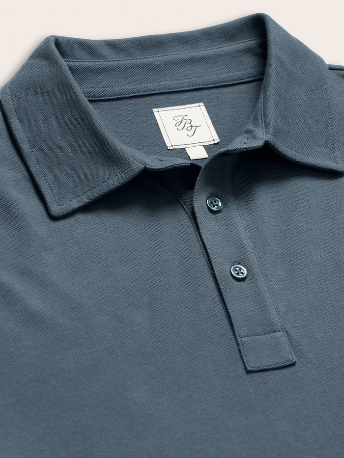 Close-up of slate blue pique polo collar and buttons with brand tag, on cream background