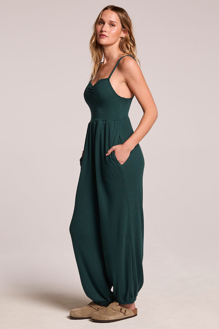 Side view of a woman wearing the Chaser  Ellery Pine Green Jumpsuit on a neutral background with hands in her pockets