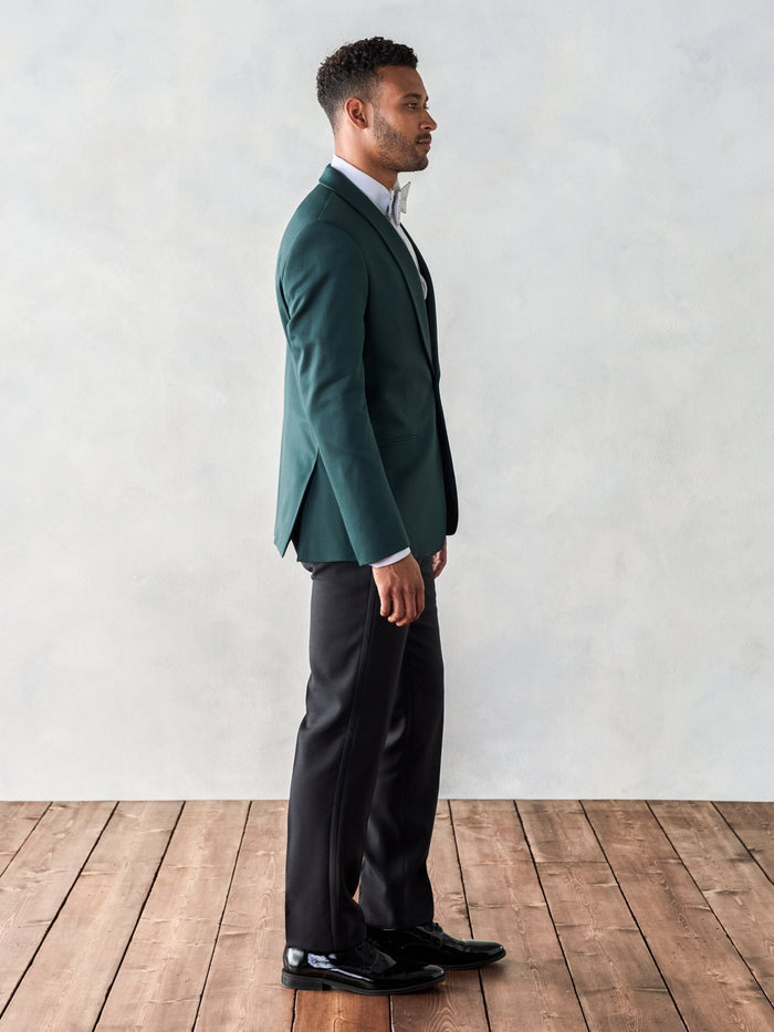 Side profile of a man standing in an emerald green shawl-lapel tuxedo jacket and black trousers.