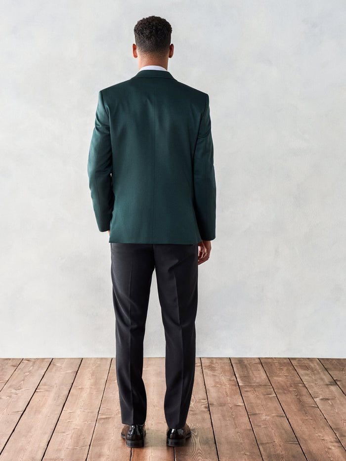 Back view of a man standing in an emerald green shawl-lapel tuxedo jacket and black trousers.