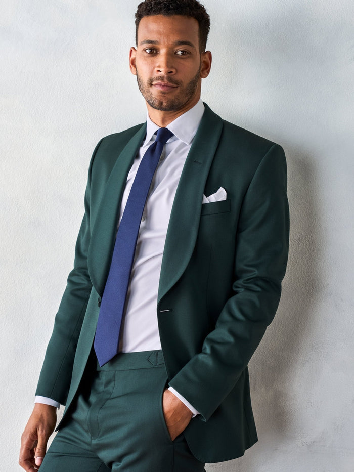 Man in an emerald green tuxedo jacket with a navy tie and white pocket square, leaning against a wall.