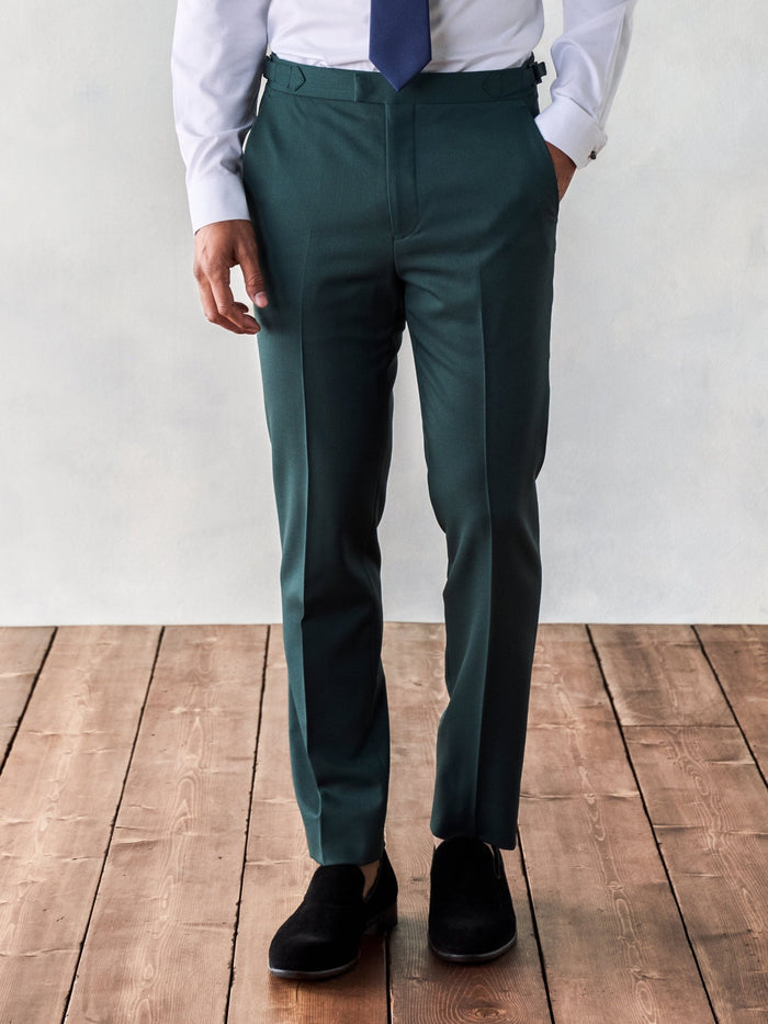 Man from waist down wearing emerald green trousers with black velvet loafers, on a wood floor.