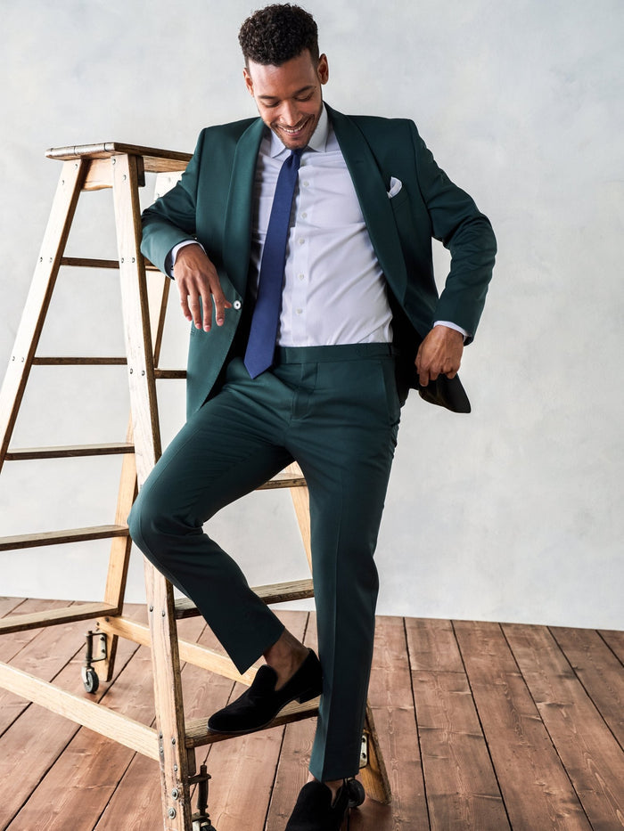 Man in an emerald green suit posing playfully on a wooden ladder, navy tie and pocket square.