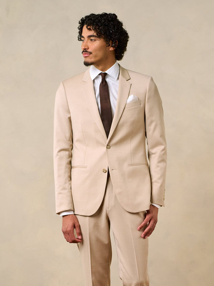 Man in a beige suit with a brown tie and white shirt, standing against a neutral background.