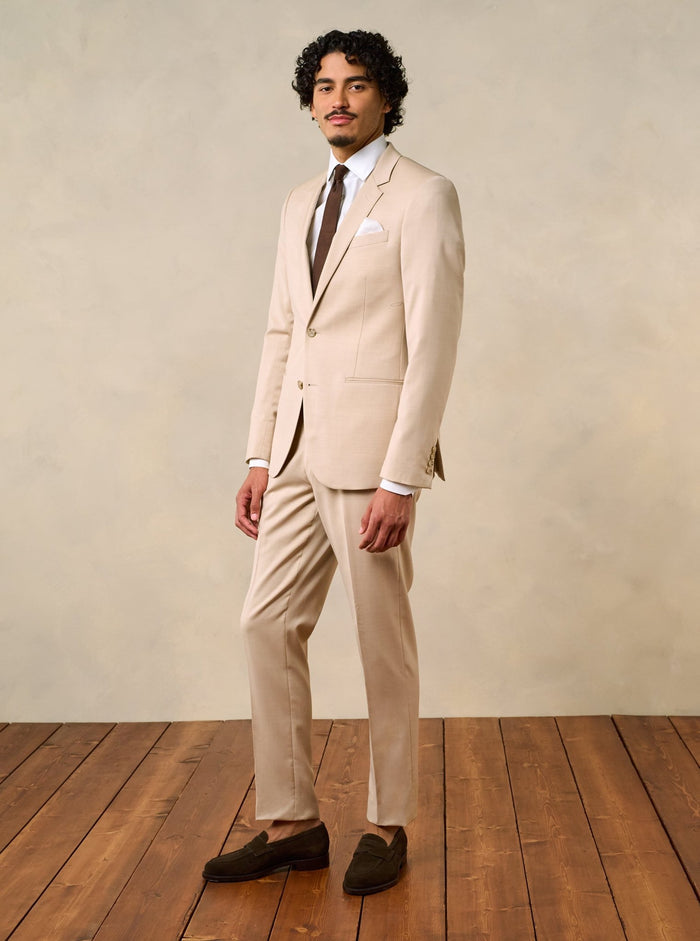 Man wearing a beige suit with a brown tie and brown shoes, standing on wooden floor.