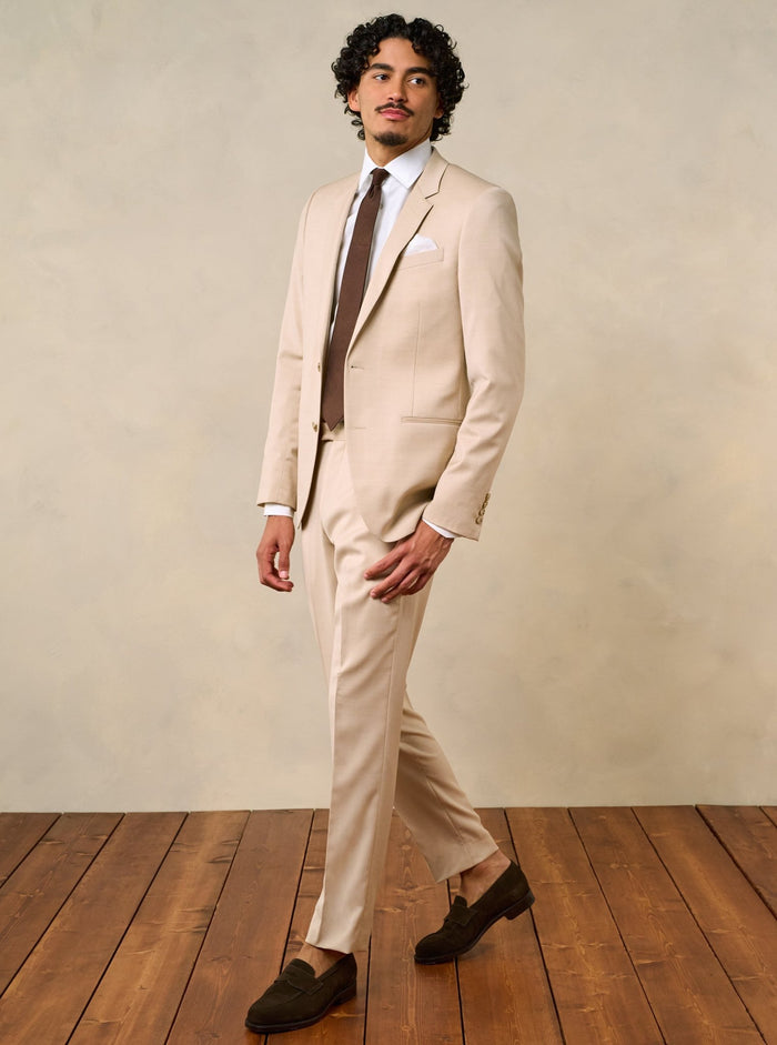 Man walking in a beige suit with a brown tie and white pocket square.