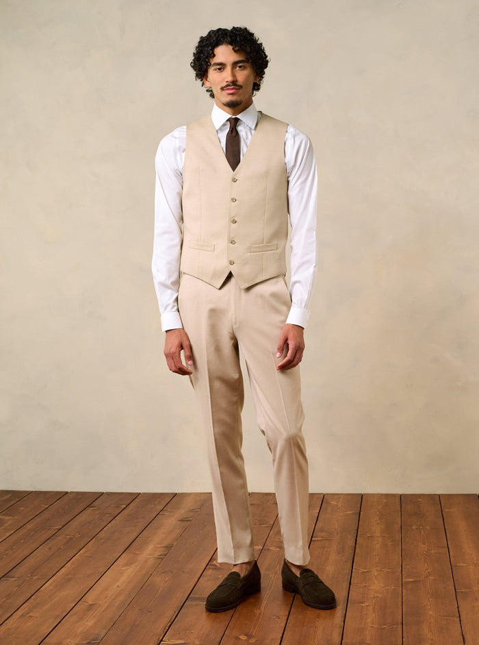 Man wearing a beige vest and pants with a white shirt and brown tie, standing on wooden floor.