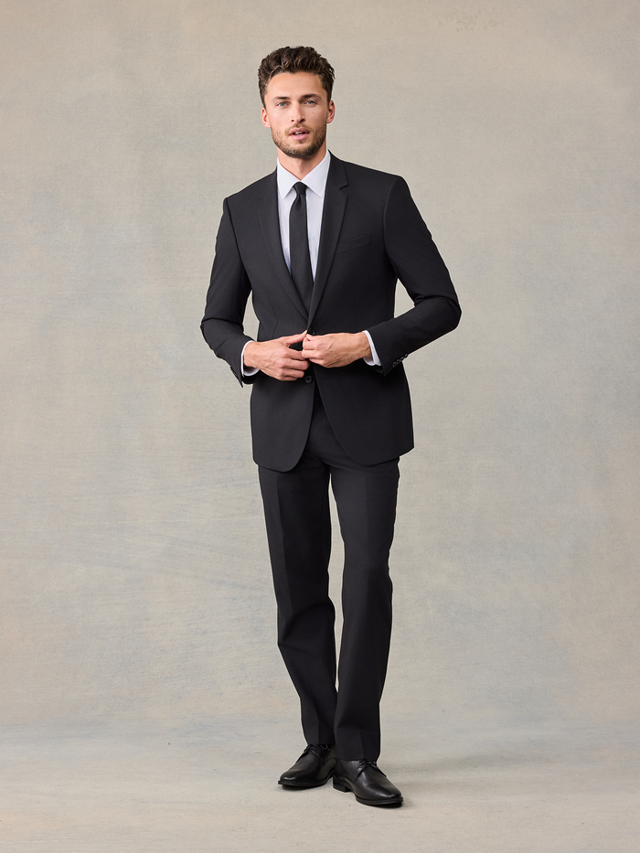 Man in a black suit buttoning his jacket, full length, with a black tie and white shirt.