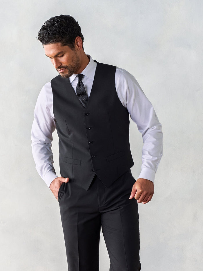 Man in a black suit vest with a patterned tie and matching trousers, looking down.