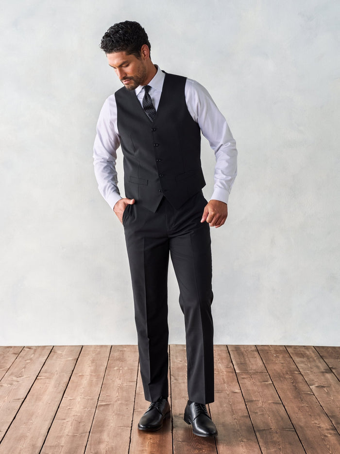 Man walking in a black suit vest with a patterned tie, on a wood floor.