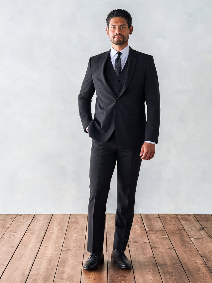Man in a black three-piece suit with a patterned tie, standing on a wood floor.