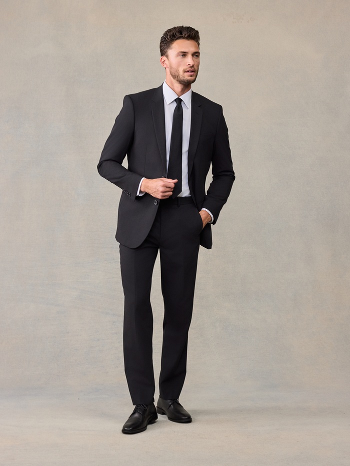 Man walking in a black suit with a black tie, adjusting his cuff.