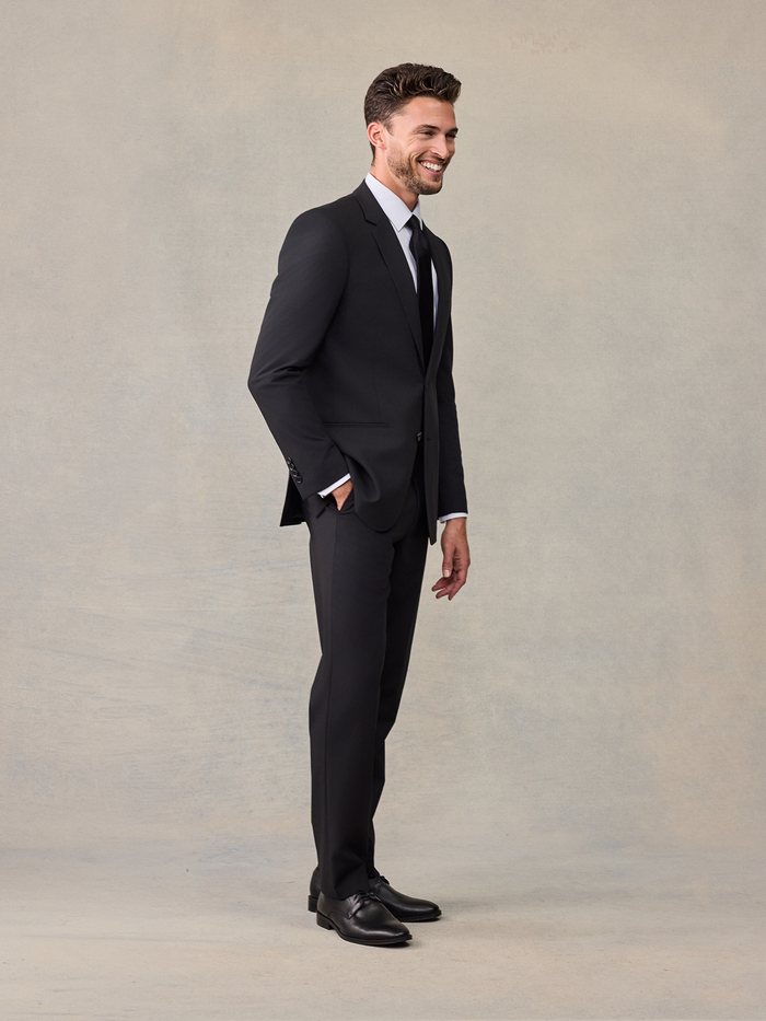 Side view of a man in a black suit, smiling, with one hand in his trouser pocket.