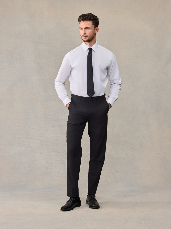 Man in a white dress shirt and black tie with black trousers, hands in pockets, no jacket.