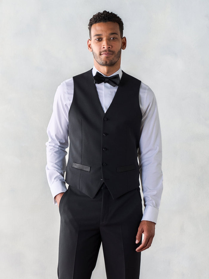 Man in a black tuxedo vest with a black bow tie and matching trousers.