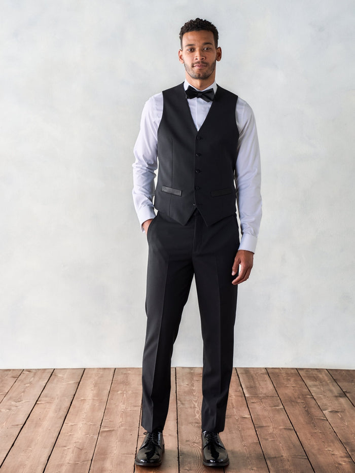 Man in a black tuxedo vest and bow tie, standing on a wood floor.