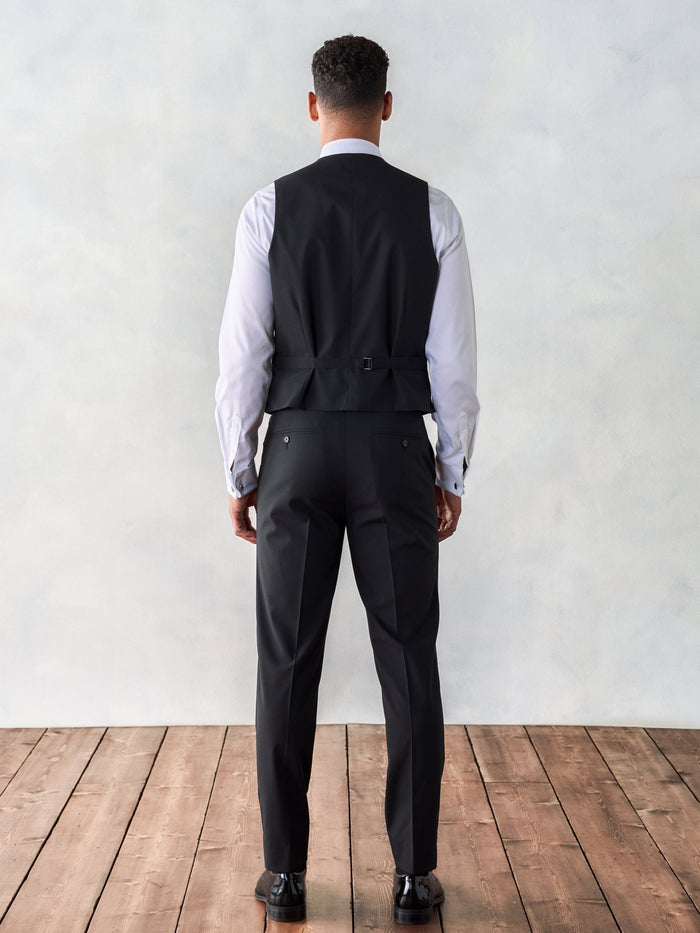 Back view of a man in a black tuxedo vest and trousers, on a wood floor.